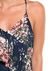 [Color: Navy/Blush/Olive] A detail image of a brunette model wearing a navy blue floral tank top with a flowy ruffle front and tie back detail. 