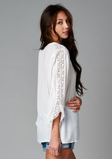 [Color: Off White] A side facing image of a brunette model wearing a bohemian white v-neck blouse with lace crochet detail at the sleeve.
