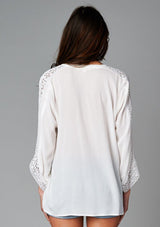 [Color: Off White] A back facing image of a brunette model wearing a bohemian white v-neck blouse with lace crochet detail at the sleeve.