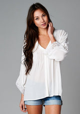 [Color: Off White] A front facing image of a brunette model wearing a bohemian white v-neck blouse with lace crochet detail at the sleeve.