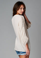 [Color: Champagne] A side facing image of a brunette model wearing a bohemian v-neck blouse with lace crochet detail at the sleeve.