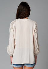 [Color: Champagne] A back facing image of a brunette model wearing a bohemian v-neck blouse with lace crochet detail at the sleeve.