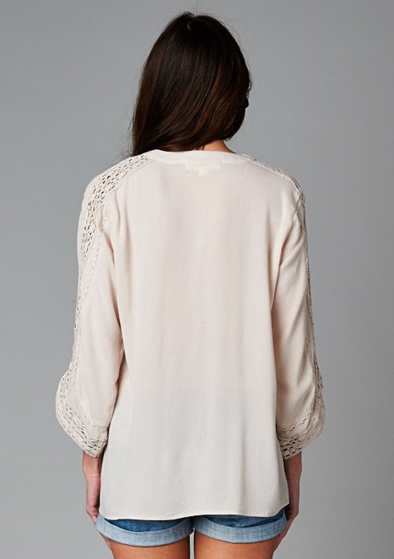 [Color: Champagne] A back facing image of a brunette model wearing a bohemian v-neck blouse with lace crochet detail at the sleeve.