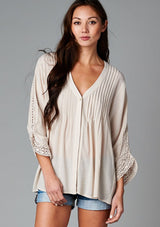 [Color: Champagne] A front facing image of a brunette model wearing a bohemian v-neck blouse with lace crochet detail at the sleeve.