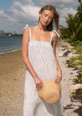 [Color: White/Denim Blue] A front facing image of a blonde model at the beach wearing a white and blue shirred maxi dress with tie shoulders.  