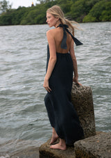 [Color: Slate Blue] A back facing image of a blonde model standing near water wearing a relaxed crinkled texture maxi dress with an o ring and halter neckline. 