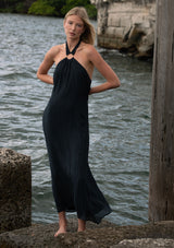 [Color: Slate Blue] A front facing image of a blonde model standing near water wearing a relaxed crinkled texture maxi dress with an o ring and halter neckline. 