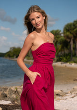 [Color: Magenta] A front facing image of a blonde model at the beach wearing a pink strapless midi dress with a gathered bust, side pockets, and a smocked back.