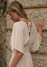 [Color: Natural/Coral] A back facing image of blonde model against a stone wall wearing a kimono sleeve maxi dress.