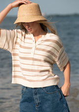 [Color: Vanilla/Taupe] A front facing image of a blonde model next to water wearing an ivory and beige cotton striped sweater top with blue denim shorts.