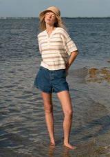 [Color: Vanilla/Taupe] A front facing image of a blonde model standing on water wearing an ivory and beige cotton striped sweater top with blue denim shorts.