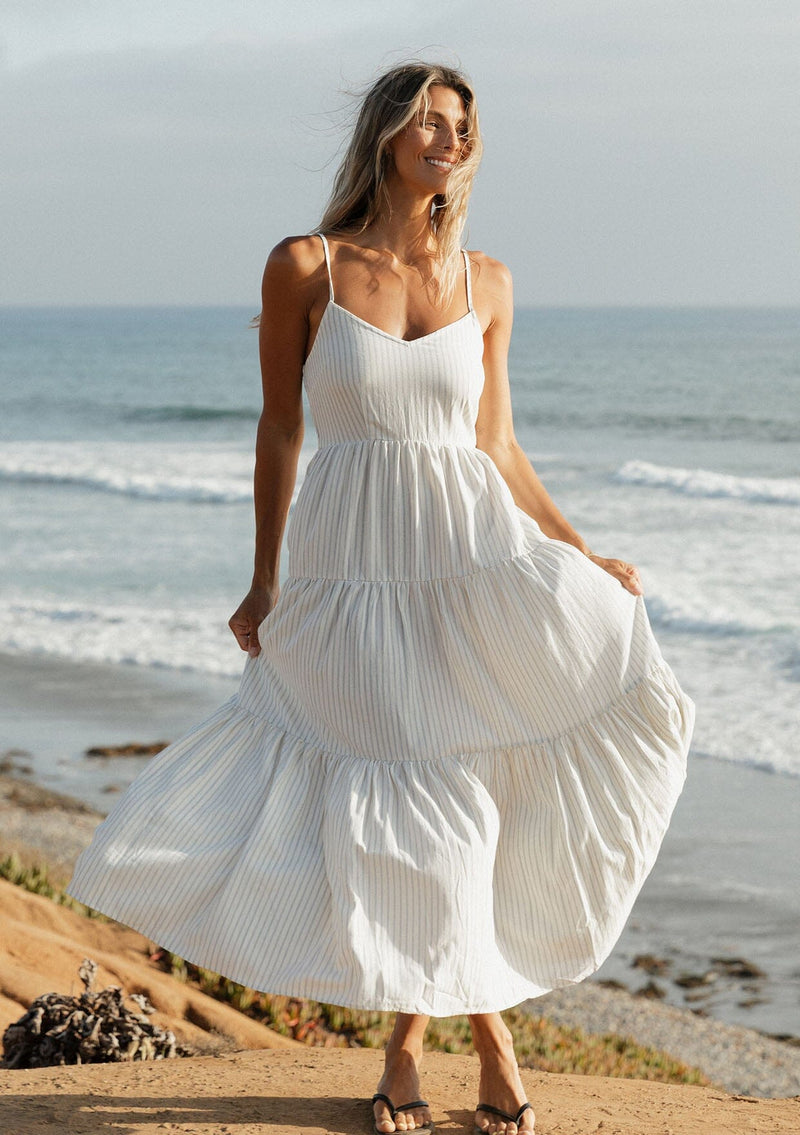 [Color: Natural/Blue] A front image of a blonde model wearing a striped maxi dress with a tiered skirt, v-neckline, adjustable spaghetti straps, smocked back, and side pockets. 