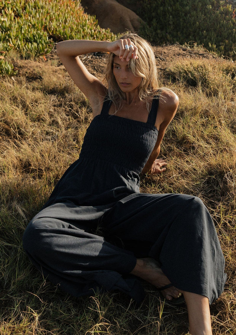 [Color: Navy] A sitting image of a blonde model at the beach wearing a navy wide leg jumpsuit with a smocked top, wide fixed straps, and side pockets. 