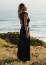 [Color: Navy] A side facing image of a blonde model wearing a navy wide leg jumpsuit with a smocked top, wide fixed straps, and side pockets. 