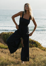[Color: Navy] A front facing image of a blonde model at the beach wearing a navy wide leg jumpsuit with a smocked top, wide fixed straps, and side pockets. 