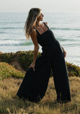 [Color: Navy] A front facing image of a blonde model at the beach wearing a navy wide leg jumpsuit with a smocked top, wide fixed straps, and side pockets. 