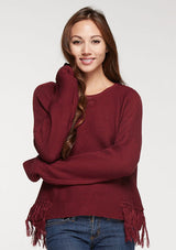 [Color: Merlot] A front facing image of a brunette model wearing a burgundy knit pullover with side fringe details, long sleeves with dropped shoulders, and a round neckline. 