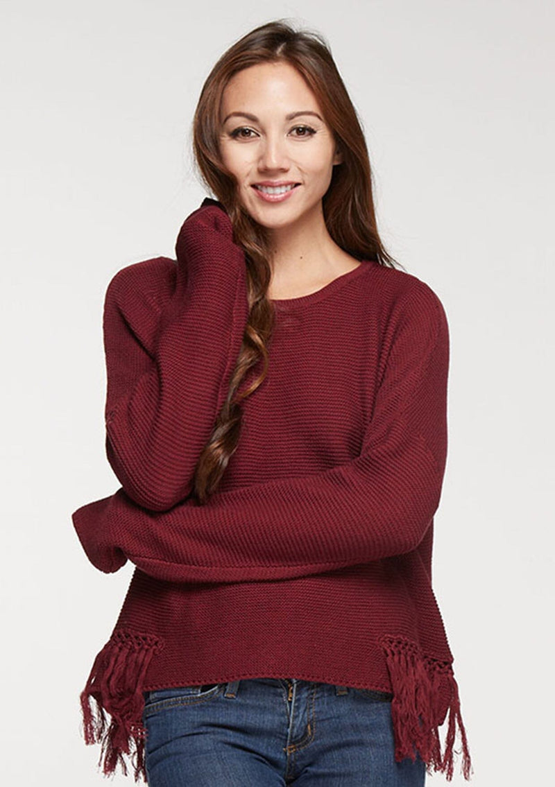 [Color: Merlot] A front facing image of a brunette model wearing a burgundy knit pullover with side fringe details, long sleeves with dropped shoulders, and a round neckline. 