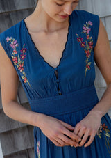 [Color: Navy] A detail image of a blonde model wearing a blue embroidered maxi dress with w scalloped v-neckline, a smocked waist, and slits. A flowy summer dress for garden parties and soirees. 