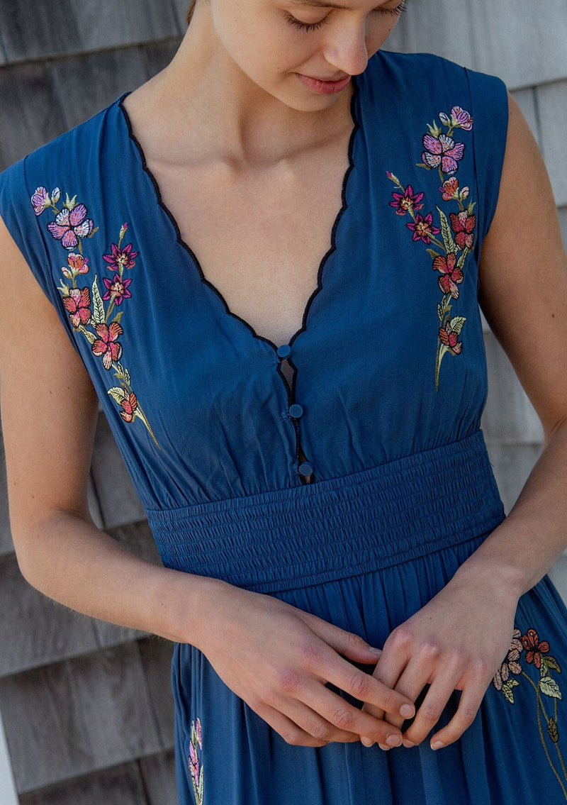 [Color: Navy] A detail image of a blonde model wearing a blue embroidered maxi dress with w scalloped v-neckline, a smocked waist, and slits. A flowy summer dress for garden parties and soirees. 