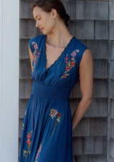 [Color: Navy] A detail image of a blonde model wearing a blue embroidered maxi dress with w scalloped v-neckline, a smocked waist, and slits. A flowy summer dress for garden parties and soirees. 