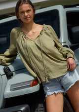 [Color: Olive] A blonde model wearing a classic lightweight sheer green peasant top. With a ruffled neckline, tassel ties, long voluminous bishop sleeves, and a relaxed flowy fit.