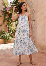 [Color: Ocean/Blue] A brunette model standing near greenery and a stone wall wearing a blue and white halter maxi dress with a relaxed, flowy silhouette. 