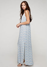[Color: Blue/Sky] A side facing image of a brunette model wearing a sleeveless maxi dress with an abstract print, a white chevron pattern, side slits, and a crochet back panel.