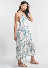 [Color: Ocean/Blue] A side facing image of a brunette model wearing a blue and white floral sleeveless halter maxi dress with a relaxed, flowy silhouette and tassel ties. Styled with brown strappy sandals. 
