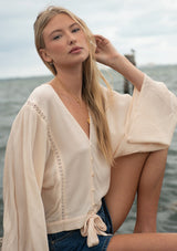 [Color: Tea Dye] A sitting image of a blonde model at the beach wearing a light beige blouse with wide long sleeves, a v neckline, button front, and a tie front detail. 