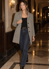 [Color: Cream/Black] A woman walking down a grand, warmly lit hotel hallway wearing a black and beige herringbone patterned blazer over a black top and dark blue jeans.