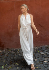 [Color: Natural] A side facing image of a blonde model against a textured red wall wearing a beige bohemian sleeveless maxi dress with a button front top, waist belt, side pockets, and contrast stitching. 