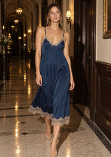[Color: Navy/Taupe] A woman walks down a grand, warmly lit hallway wearing a navy blue satin slip dress with beige lace trim along the straps and hem, paired with nude heels.
