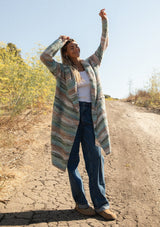 [Color: Mint/Natural] A front facing image with a blonde model wearing a cozy bohemian space dye chunky knit cardigan in a green mint multi-color stripe pattern. This plush cardigan features long sleeves, a cozy hood, essential pockets, and an open front. The long length and thick knit make it the perfect sweater for the fall season.