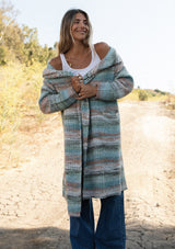 [Color: Mint/Natural] A front facing image with a blonde model wearing a cozy bohemian space dye chunky knit cardigan in a green mint multi-color stripe pattern. This plush cardigan features long sleeves, a cozy hood, essential pockets, and an open front. The long length and thick knit make it the perfect sweater for the fall season.