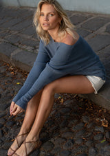[Color: Heather Ocean] A sitting image of a blonde model wearing a blue waffle knit pullover sweater. With long sleeves, a relaxed fit, and a wide neckline that can be worn off the shoulder.