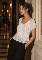 [Color: White] A woman wearing a white sheer puff-sleeve blouse with a lace hem and black trousers, leaning against a cream-colored wall in an elegant, warmly lit hallway.