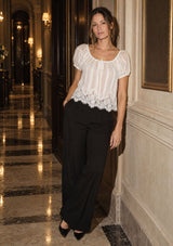 [Color: White] A woman stands in a luxurious hotel hallway wearing a white short-sleeve blouse with a sheer striped bodice, a floral lace hem, and black wide-leg trousers.