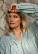 [Color: Dusty Sage] A detail image of a blonde model wearing green lightweight blouse with pintuck accents, a button front, and split v-neckline. Airy and sheer for any warm weather occasion. 