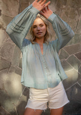 [Color: Dusty Sage] A front facing image of a blonde model wearing green lightweight blouse with pintuck accents, a button front, and split v-neckline. Airy and sheer for any warm weather occasion. 