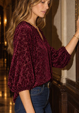 [Color: Merlot] A woman wearing a deep burgundy velvet blouse with a subtle tonal floral pattern, paired with blue jeans, posing indoors against a dark wood-paneled wall.