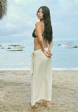 [Color: Vanilla] A back facing image of a brunette model wearing an off-white linen blend wrap skirt with a side tie and smocked back, styled for warm weather, vacation, or beach days.