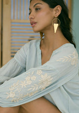 [Color: Dusty Sage/Natural] A detail image of a brunette model wearing a green boho embroidered blouse with a split v-neckline, lace trim details, long balloon sleeves, and smocked cuffs. 