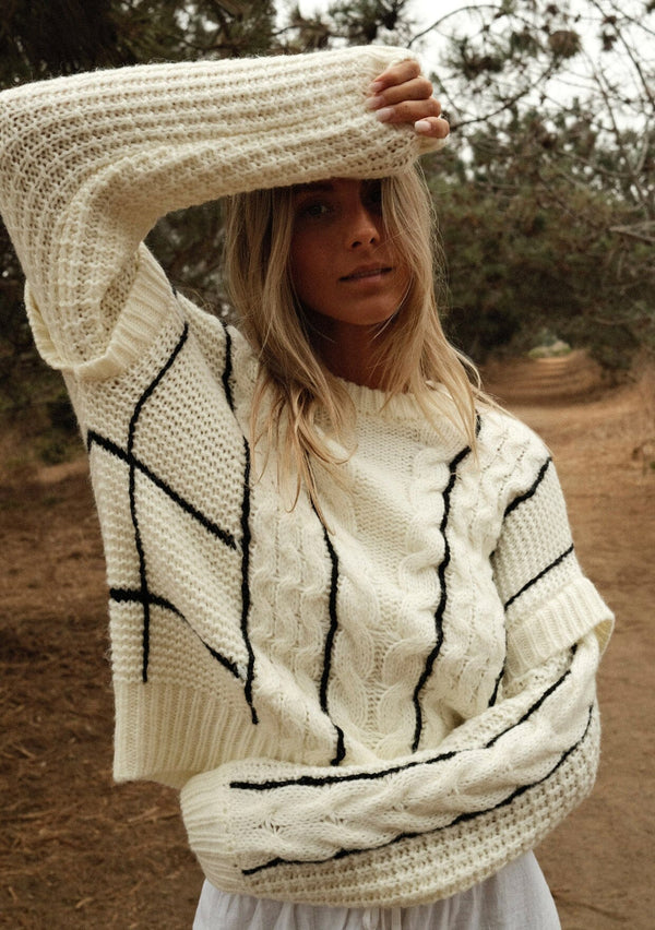 [Color: Cream/Black] A front facing image of a blonde model wearing a cozy knit sweater with a mixed fabric patchwork and cable knit detailing. This casual sweater features a crew neckline, long sleeves with a dropped shoulder, and a contrasting stripe accent. A cold weather staple for the fall and winter season. 