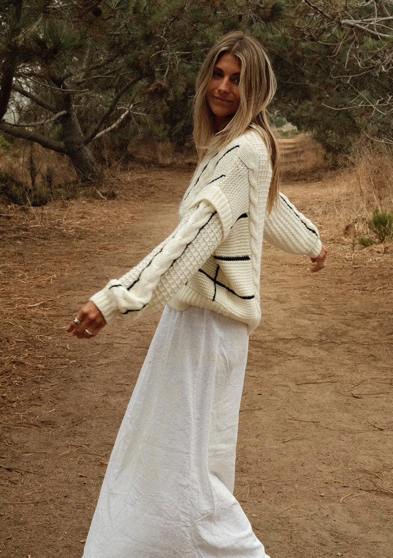 [Color: Cream/Black] A side facing image of a blonde model wearing a cozy knit sweater with a mixed fabric patchwork and cable knit detailing. This casual sweater features a crew neckline, long sleeves with a dropped shoulder, and a contrasting stripe accent. A cold weather staple for the fall and winter season. 