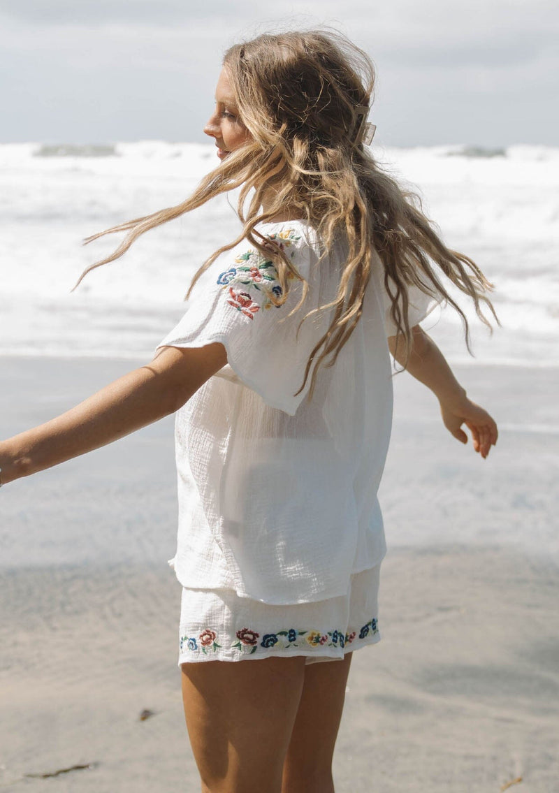 [Color: Cream] A side facing image of a blonde model wearing a light sheer cotton top with floral embroidery. Featuring a short dolman sleeves, a split v-neckline, and button front. 