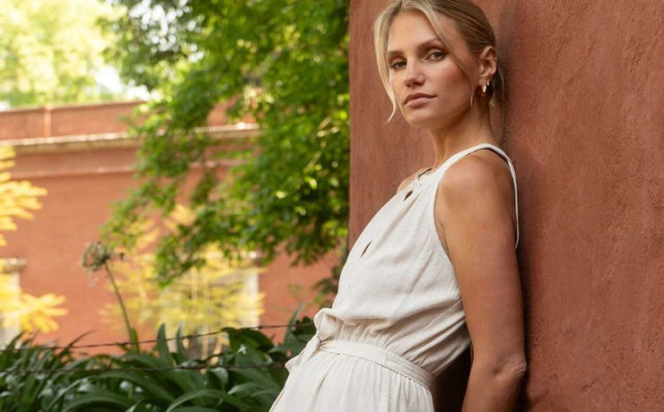 Woman in a white dress leaning against a red brick wall with greenery in the background