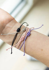 [Color: Blush] Four adjustable cord bracelets in black, silver, pink, and purple knotted styles, shown worn on a person's wrist.