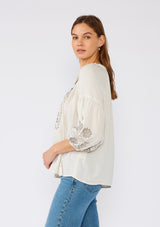 [Color: Natural/Taupe] A side facing image of a brunette model wearing an ivory flowy bohemian peasant top with embroidered detail and tassel ties.