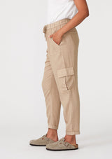 [Color: Sand] A side facing image of a brunette model wearing a khaki cargo pant with a cropped tapered leg, side pockets, an elastic waist, and a drawstring tie waist.
